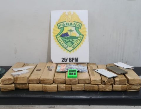  Tabletes de maconha dispostos em uma mesa, junto a uma placa com o brasão da Polícia Militar 