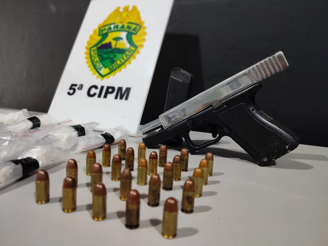  Arma, munições e drogas em cima de uma mesa, com uma placa da Polícia Militar ao fundo 