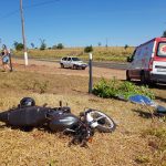 Motocicleta caída após acidente com corpo de jovem coberto ao lado e ambulância do SAMU ao fundo em uma área rural