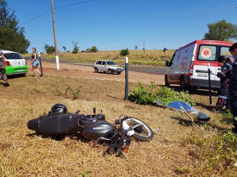  Motocicleta caída após acidente com corpo de jovem coberto ao lado e ambulância do SAMU ao fundo em uma área rural 