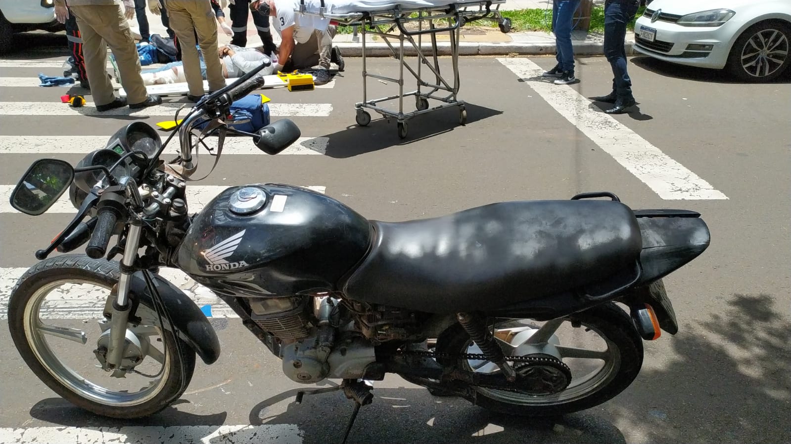  Moto com a roda dianteira destruída em primeiro plano. Homem sendo atendido pelo corpo de bombeiros em uma maca ao fundo 