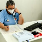 Homem sentado em uma mesa, utilizando um telefone, com o uniforme da Prefeitura de Maringá