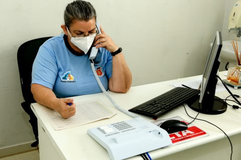  Homem sentado em uma mesa, utilizando um telefone, com o uniforme da Prefeitura de Maringá 