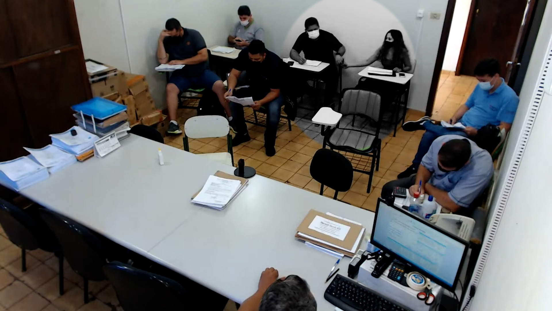  Imagem da câmera de segurança da sala de licitações. Sete pessoas sentadas em carteiras escolares, uma mesa grande e, atrás dela, o responsável pelo pregão. A imagem mostra o momento da troca de um bilhete entre dois empresários, no fundo da sala 