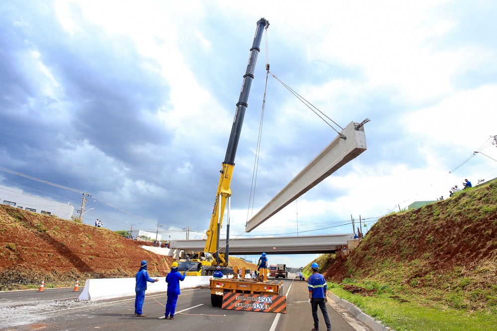  Caminhão-guindaste içando uma das vigas de concreto, presa por cabos de aço no guindaste, no meio da rodovia 