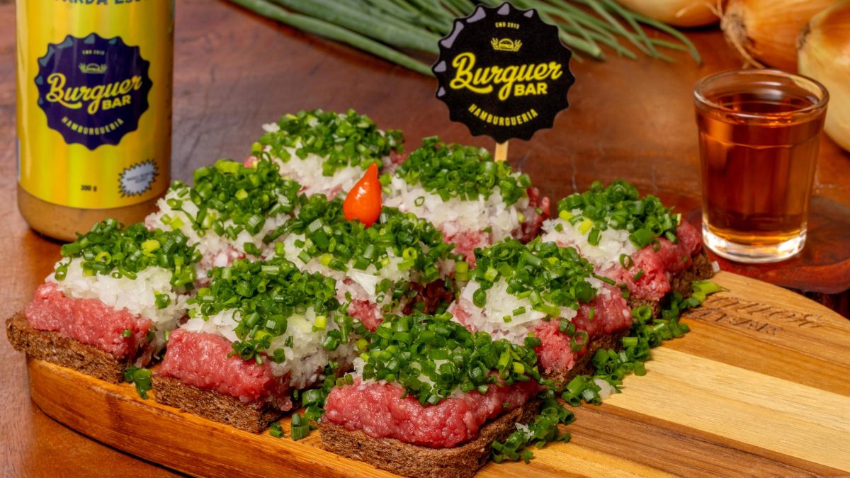 Receita do Burguer Bar leva carne bovina moída (patinho), cebola branca, cheiro verde servido na broa de centeio; acompanha mostarda escura e um shot de cachaça artesanal 